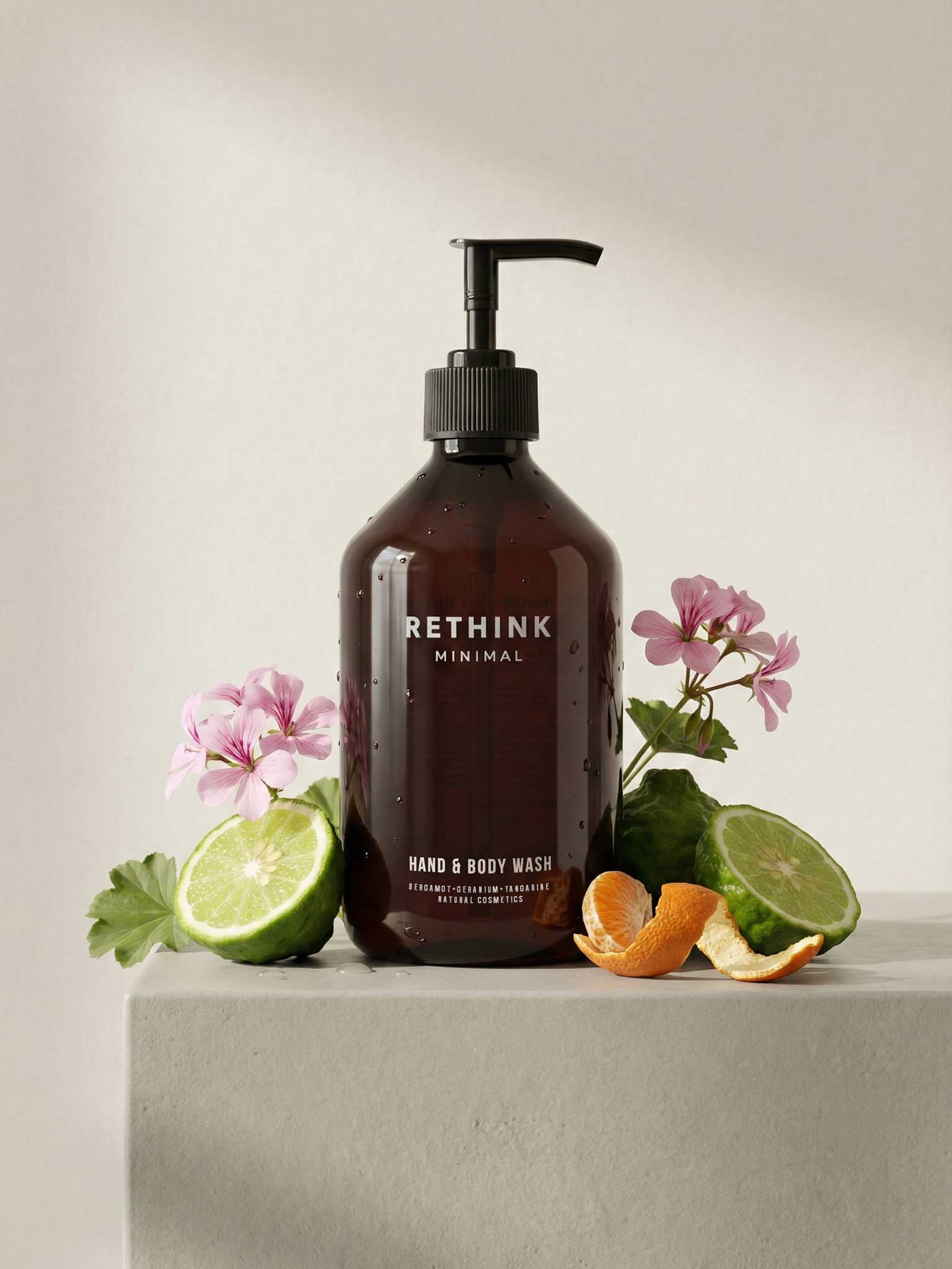 Braune Flasche für Hand- und Body Wash auf hellem Podest, umgeben von Bergamotte (Zitrusfrucht, ähnlich einer Limette), Mandarine und Geranien (Blüten) als Hinweis auf die enthaltenen natürlichen Inhaltsstoffe.